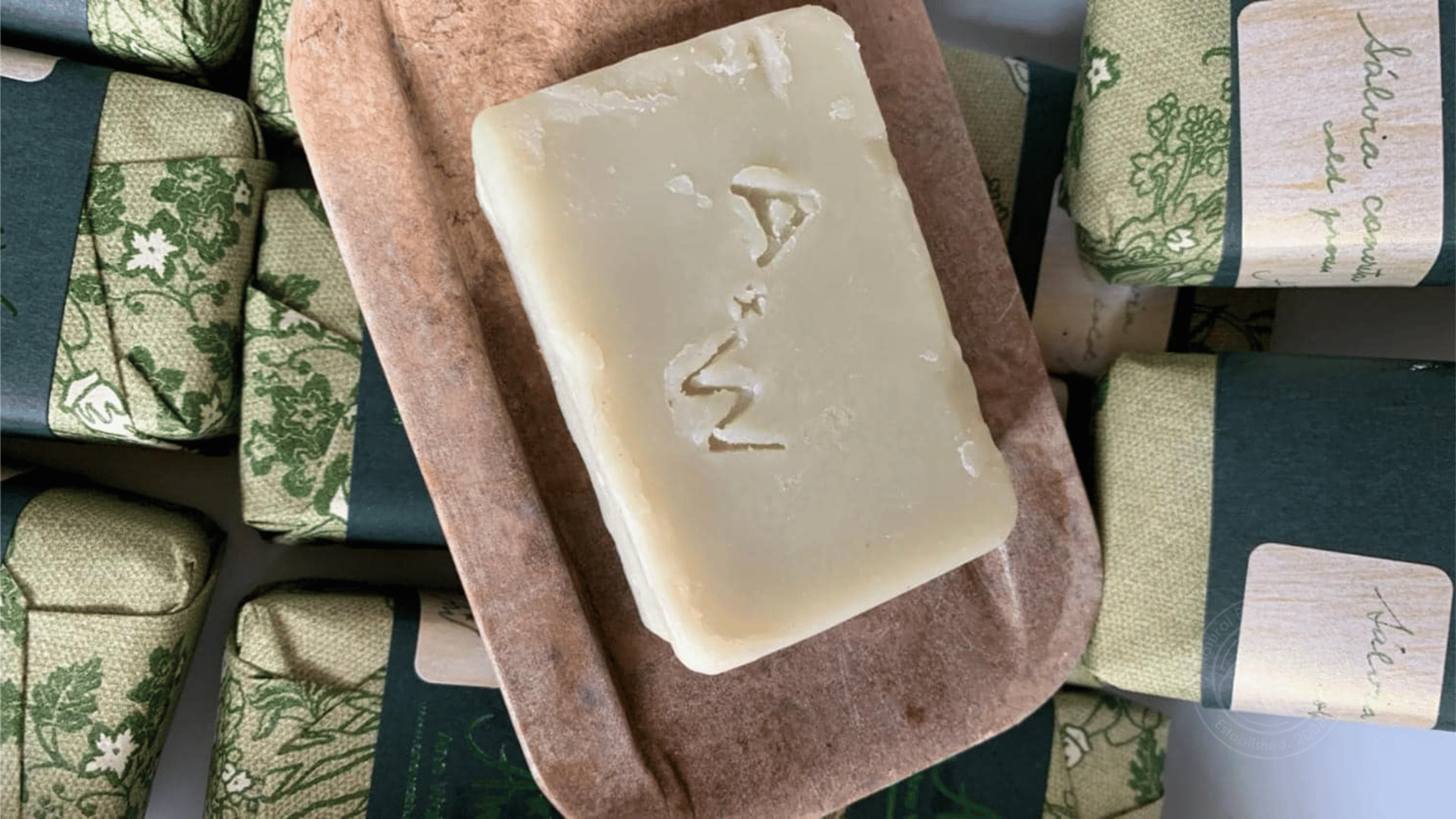 A single bar of handmade ash soap with a rustic texture, resting on a natural soap dish, wrapped in floral green paper.